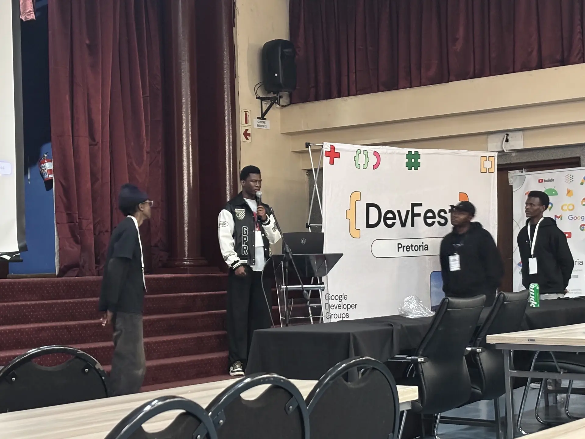 FORGE members at the front of the DevFest Pretoria hall, one speaking into a microphone while the others stand by.