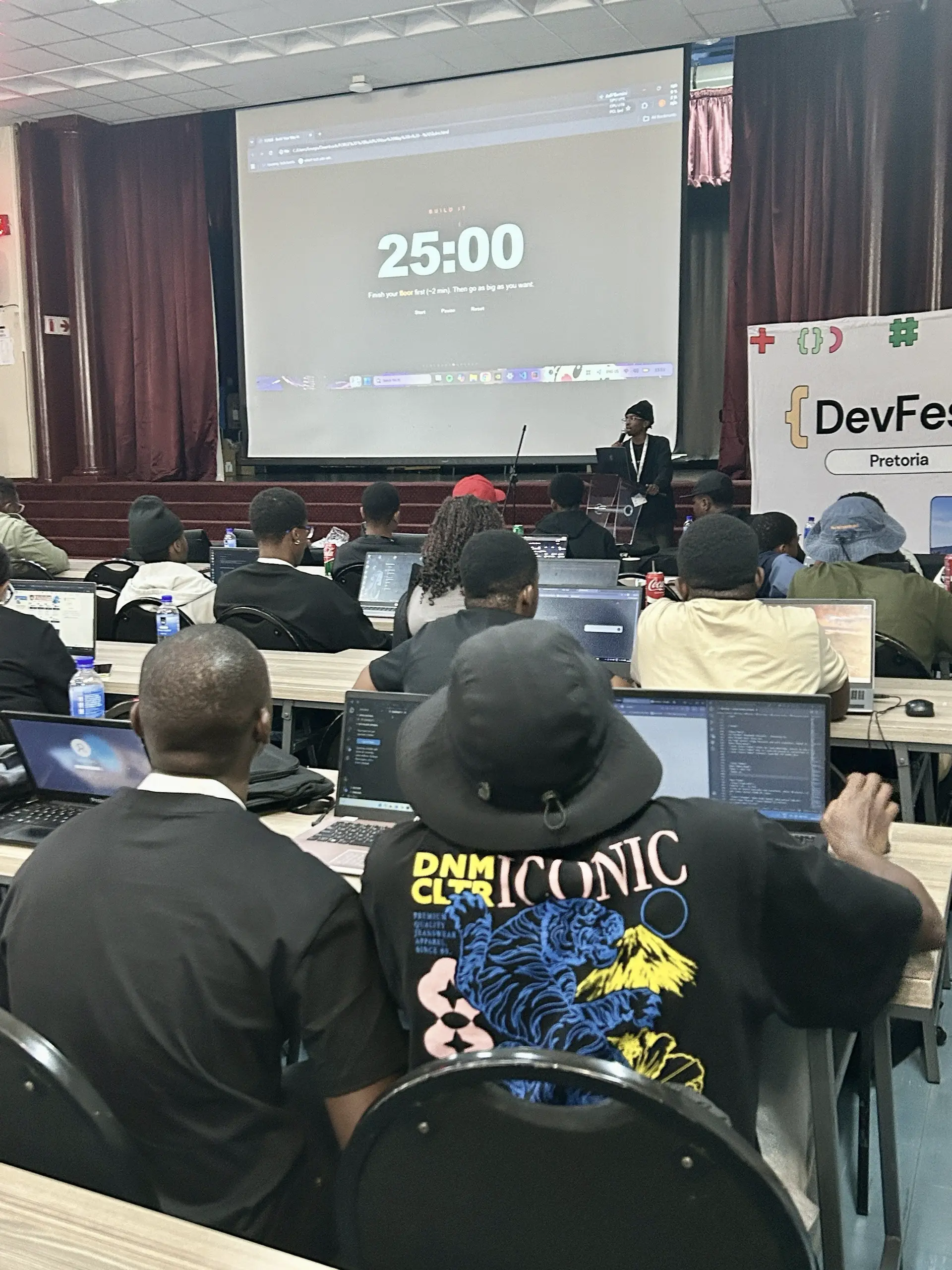 The FORGE session at DevFest Pretoria: a FORGE member presents while rows of developers code on laptops, with a 25-minute “Build it” timer on the big screen.